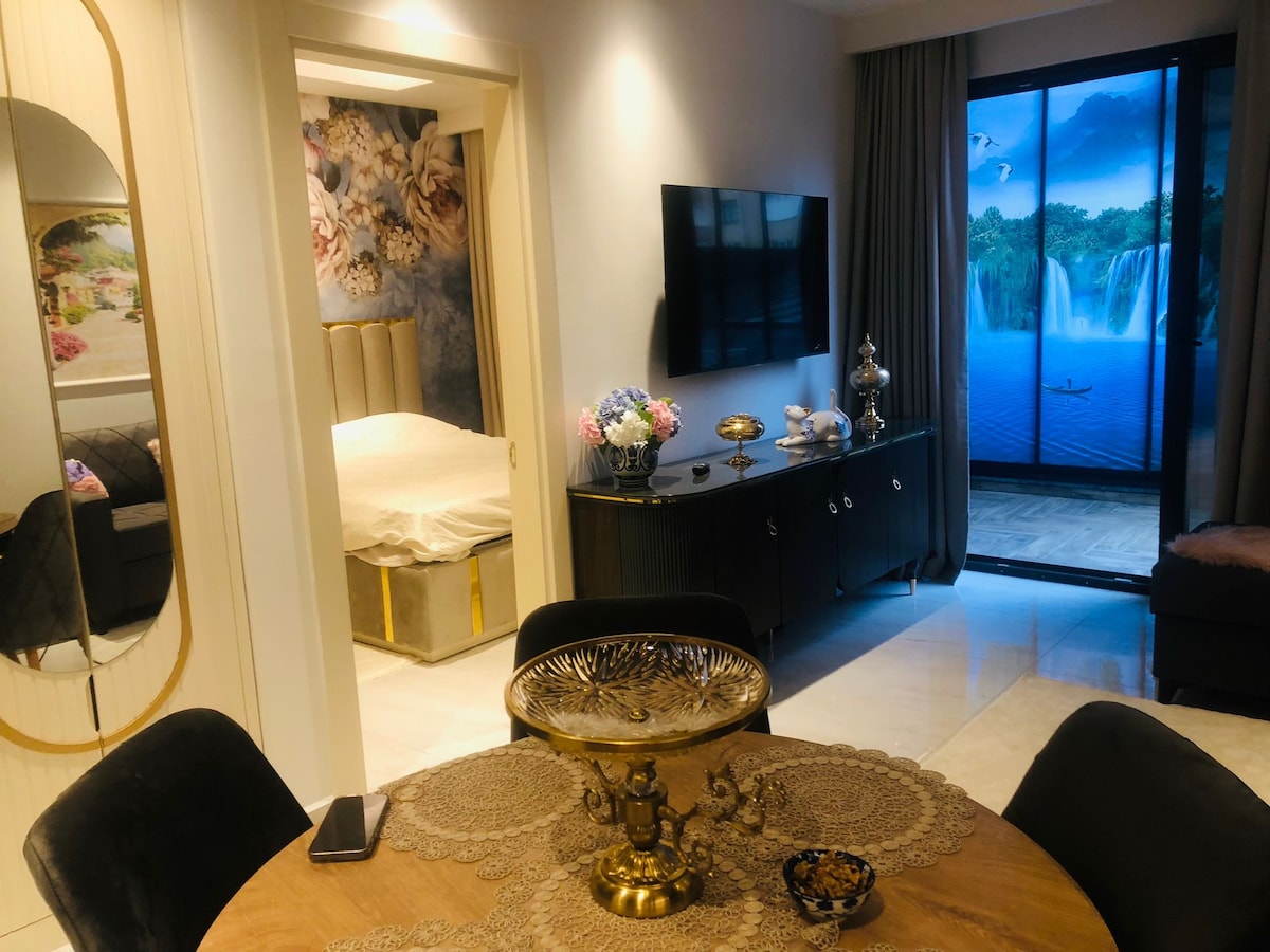 A modern living space features a round dining table with an intricate tablecloth. A black television unit sits against a wall with floral artwork, while a cozy sleeping area is visible beyond. Large glass doors reveal a serene outdoor view with a waterfall and waterway.