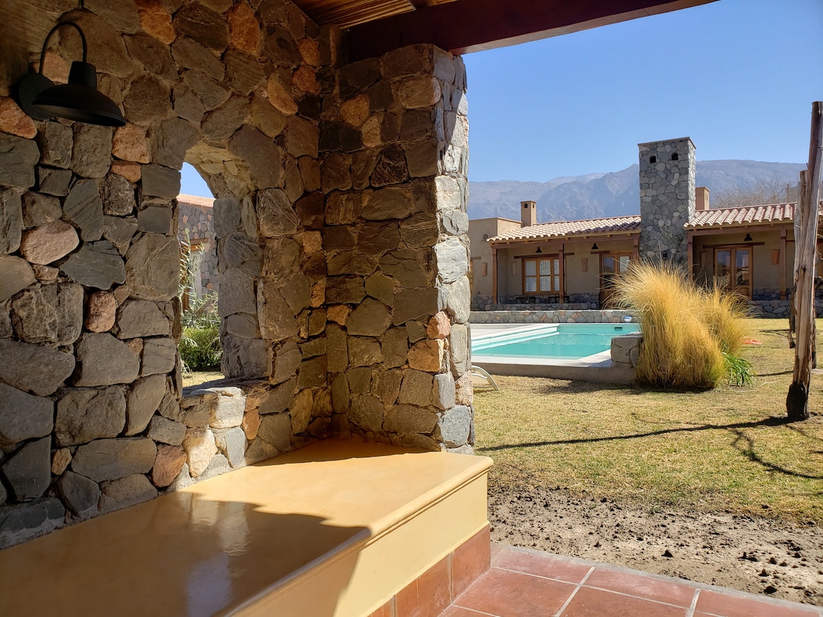 An outdoor view showcases a stone archway leading to a swimming pool surrounded by landscaped grass. The background features a single-story house with a rustic design, while mountains rise in the distance under a clear blue sky.