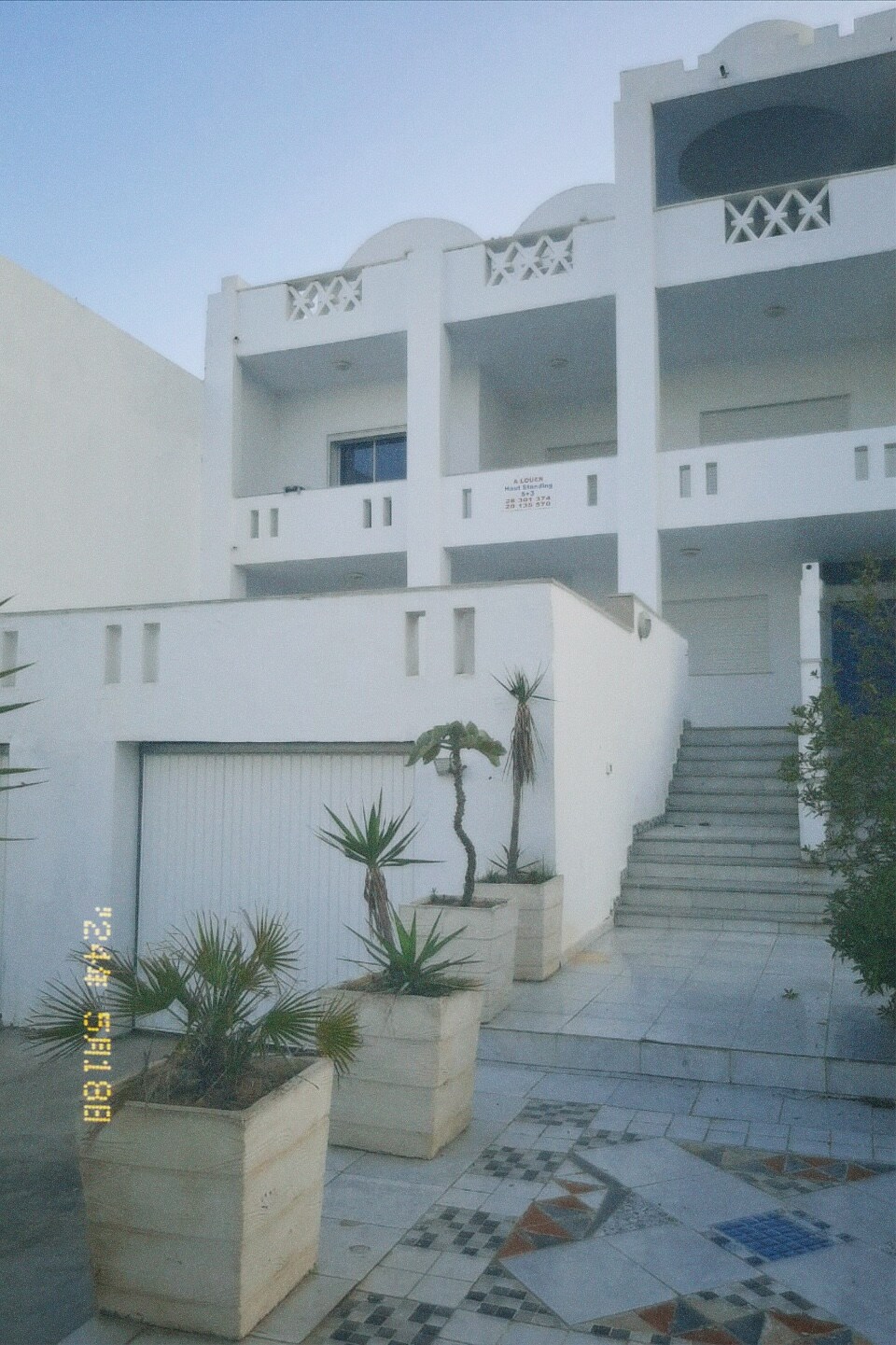 The exterior of a white multi-story building is highlighted, featuring a set of stairs leading to an entrance. Potted plants are arranged along the entryway, complementing the clean lines of the architecture. A garage door is visible at the base of the structure.