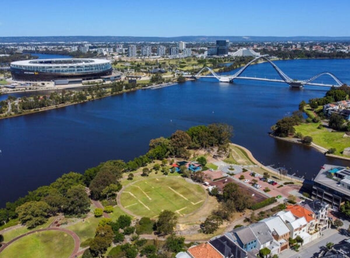 An aerial view captures the expansive Swan River, with modern architecture lining the banks. Optus Stadium is prominently visible to the left, while the striking Trafalgar Bridge arches gracefully over the water. Green spaces and pathways offer recreational areas along the river's edge.