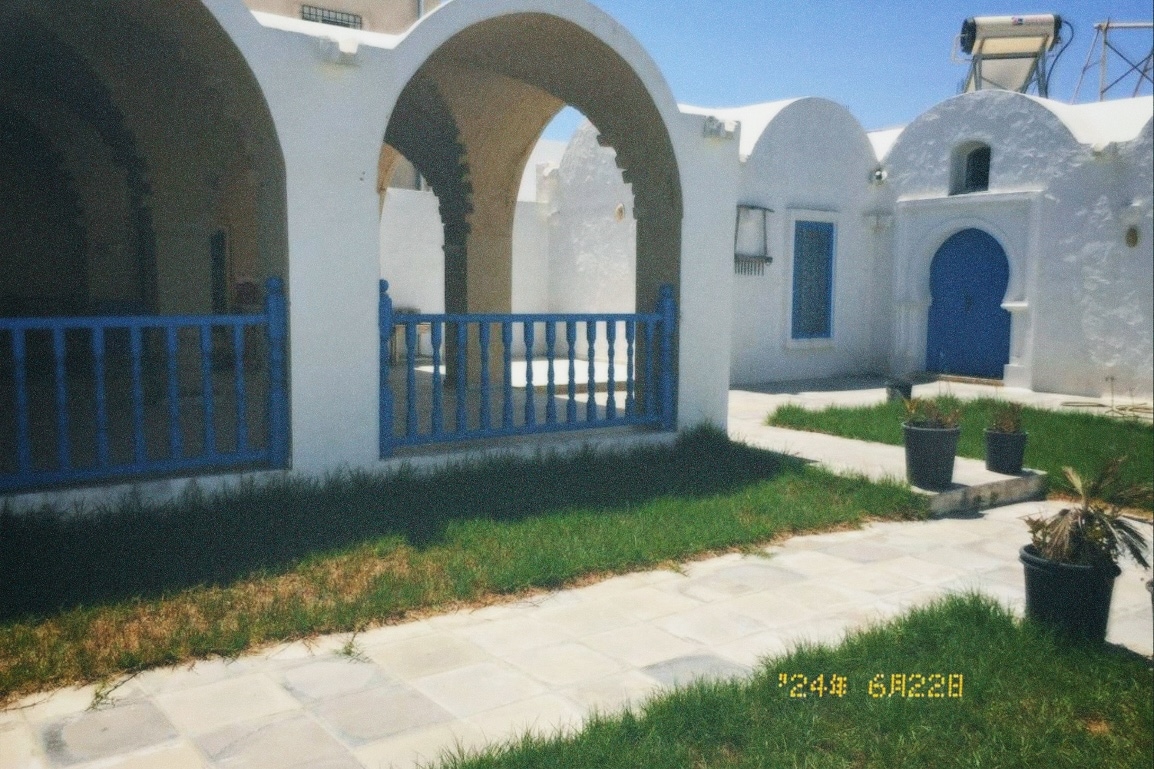 An outdoor area features whitewashed arches and blue railing, surrounded by lush green grass. Stone tiles create pathways, leading to an arched entrance that is partially visible. Potted plants line the space, contributing to a serene and inviting setting.