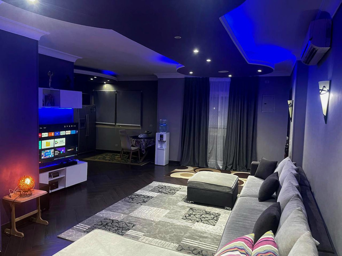 A modern living area is highlighted by blue ambient lighting and a plush sectional sofa. A large television is positioned near the wall, and a stylish area rug complements the space. Dark curtains frame the window, and a small dining table is visible in the background.