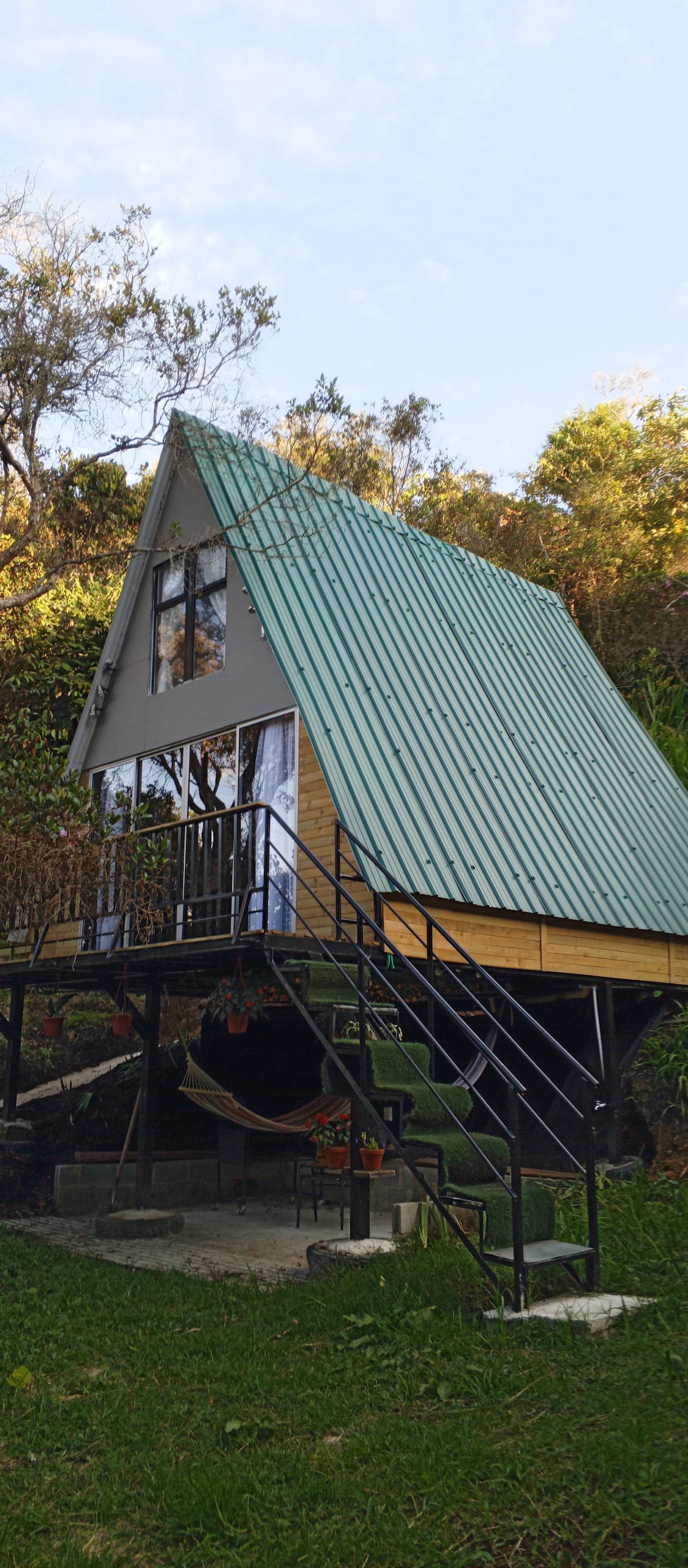 A charming A-frame cabin is situated on a grassy area, elevated on stilts. Its blue metal roof complements the surrounding greenery. Large glass windows allow natural light to fill the interior, while a staircase leads to a welcoming porch area adorned with plants.