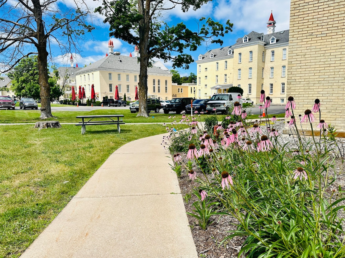 A pathway winds through a landscaped area, flanked by blooming pink flowers. In the distance, historic buildings with red-tipped roofs are visible, alongside picnic tables on a well-maintained lawn. A clear sky with scattered clouds provides a bright backdrop.