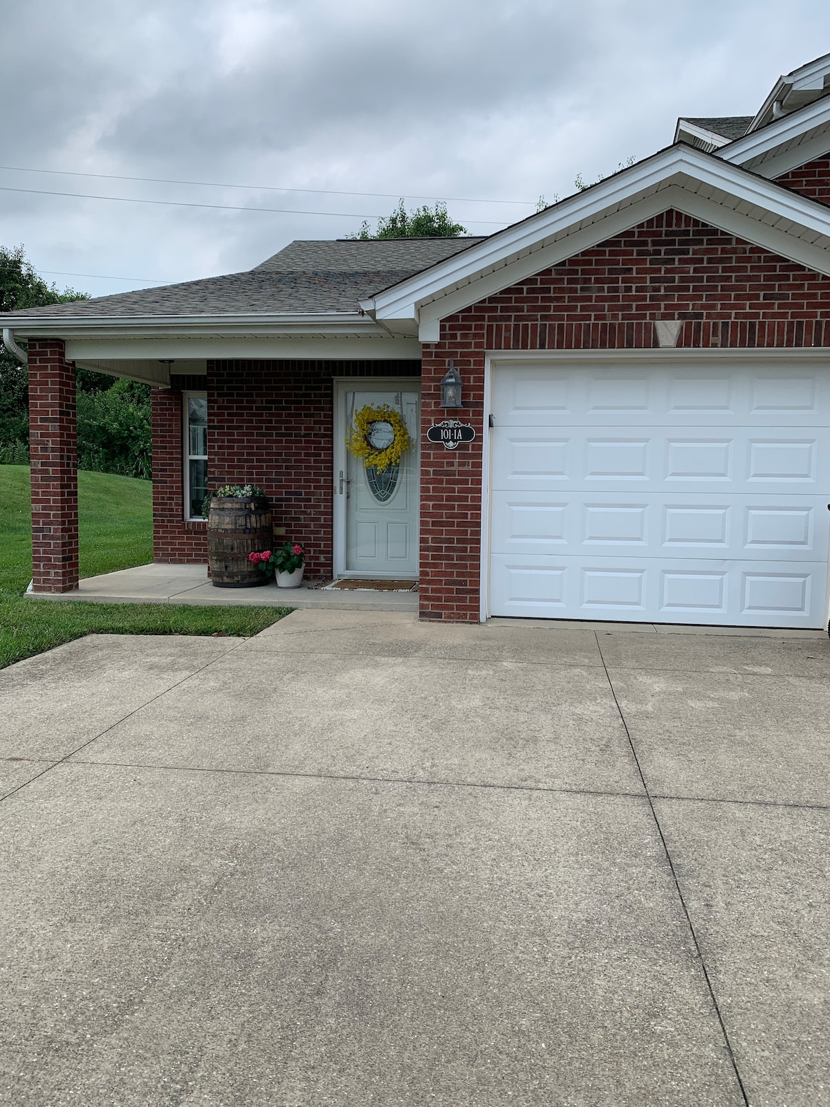 The exterior of a brick condo is shown, featuring a welcoming front door adorned with a yellow wreath. A manicured lawn and a small flower pot are visible beside the door, while a driveway leads to a white garage door.