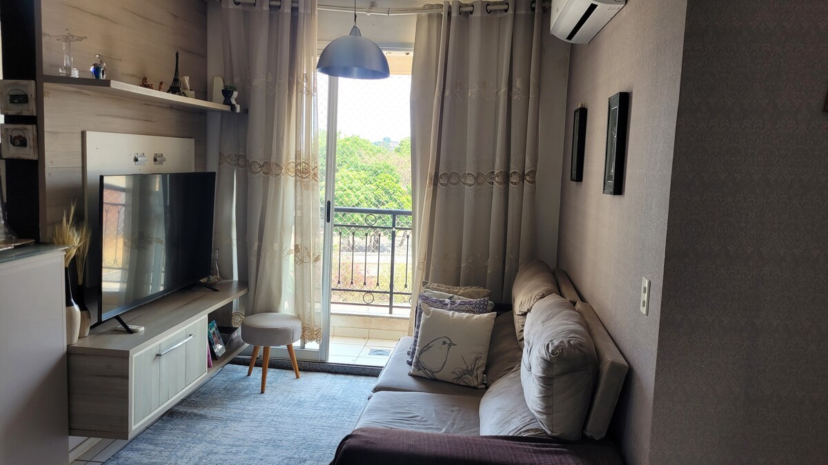 A living room area features a comfortable sofa facing a television. Light filters through sheer curtains, and a small table completes the space. A balcony is visible in the background, surrounded by greenery, enhancing the room's bright and inviting atmosphere.