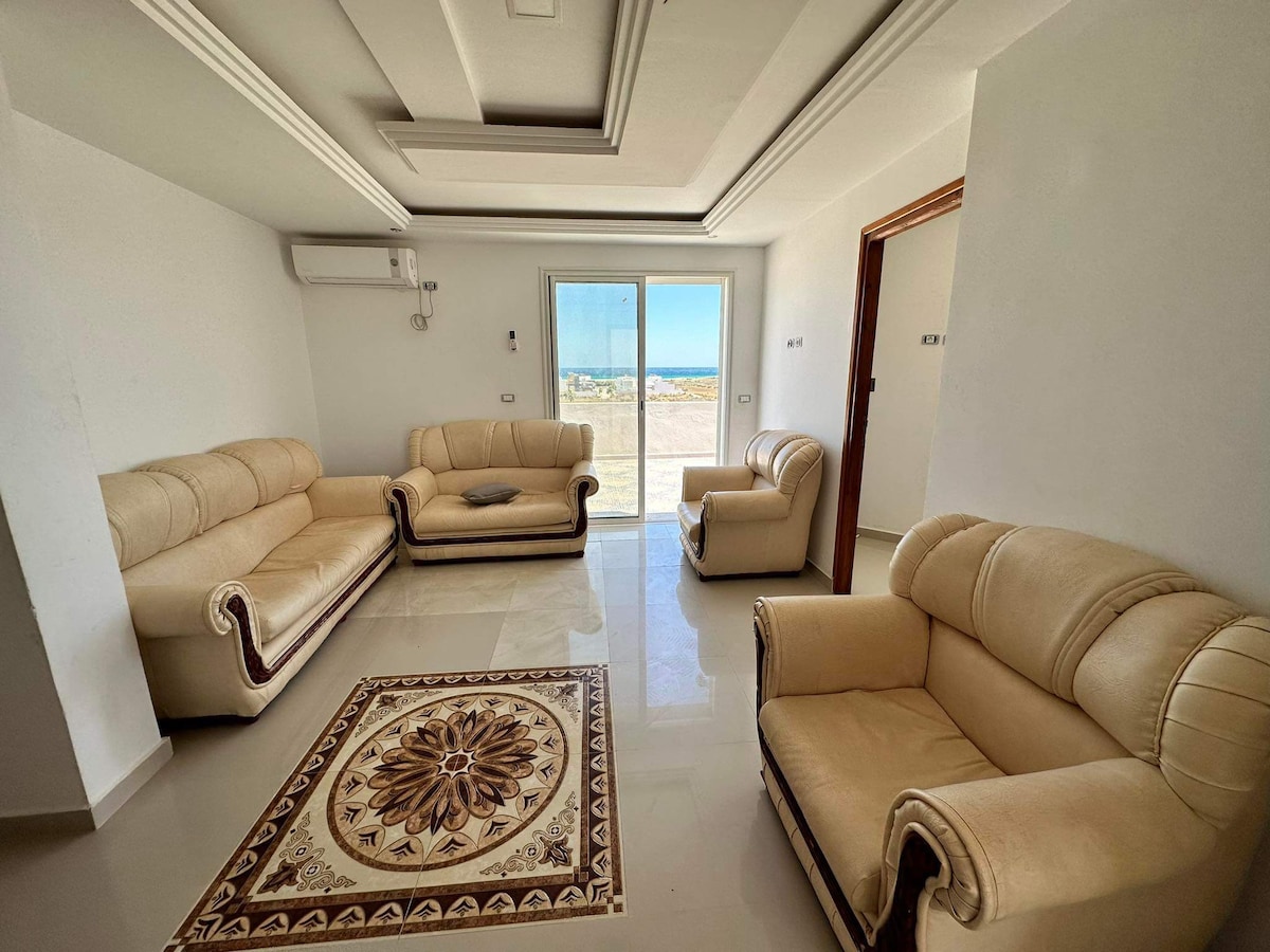 A light-filled living area features cream-colored sofas and armchairs arranged around a decorative area rug. Large sliding glass doors lead to a view of the outdoors, while light reflects off the glossy tiled floor. A neutral color palette creates a calm atmosphere.