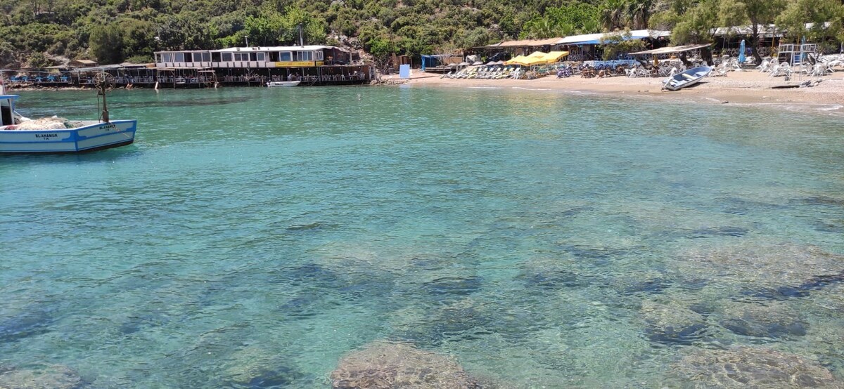 A clear view of calm, turquoise waters extends towards a sandy beach. Beachside facilities, including sun loungers and umbrellas, are visible in the background. A small boat floats gently on the water, which reflects the surrounding greenery and coastal atmosphere.