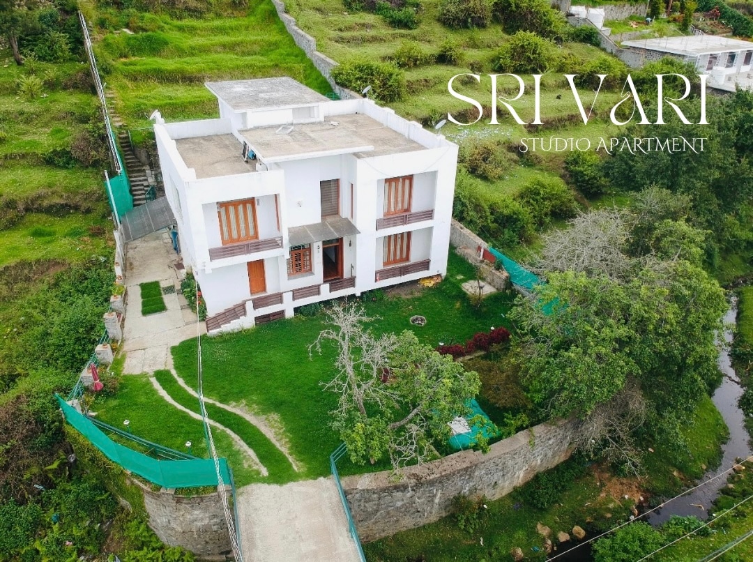 The aerial view showcases a modern studio apartment situated on a lush hillside. The spacious exterior features a well-maintained garden, greenery surrounding the building, and a pathway leading to the entrance. Large windows invite natural light into the indoor space.
