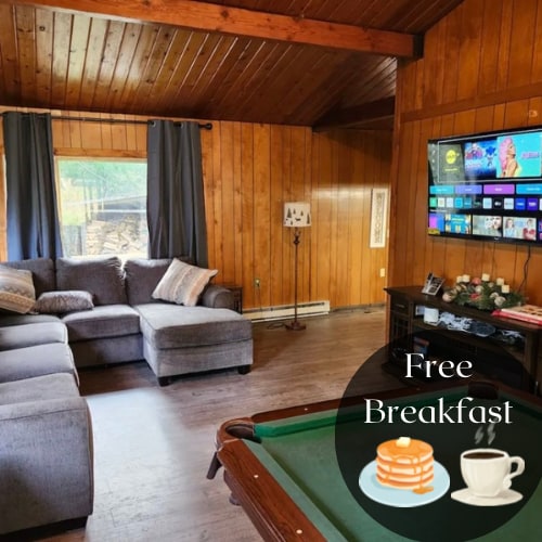 A spacious living area features a large grey sectional sofa complemented by decorative pillows. A coffee table sits in front, and a media center with a flat-screen TV is mounted on the wall. Natural light filters through large windows, illuminating the warm wooden paneling.