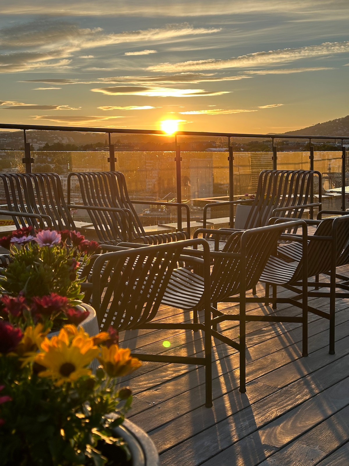 An outdoor rooftop terrace is presented, featuring several black chairs positioned to capture sunset views. Planters with vibrant flowers enhance the space, while the wooden deck reflects the warm hues of the setting sun, creating a serene atmosphere.