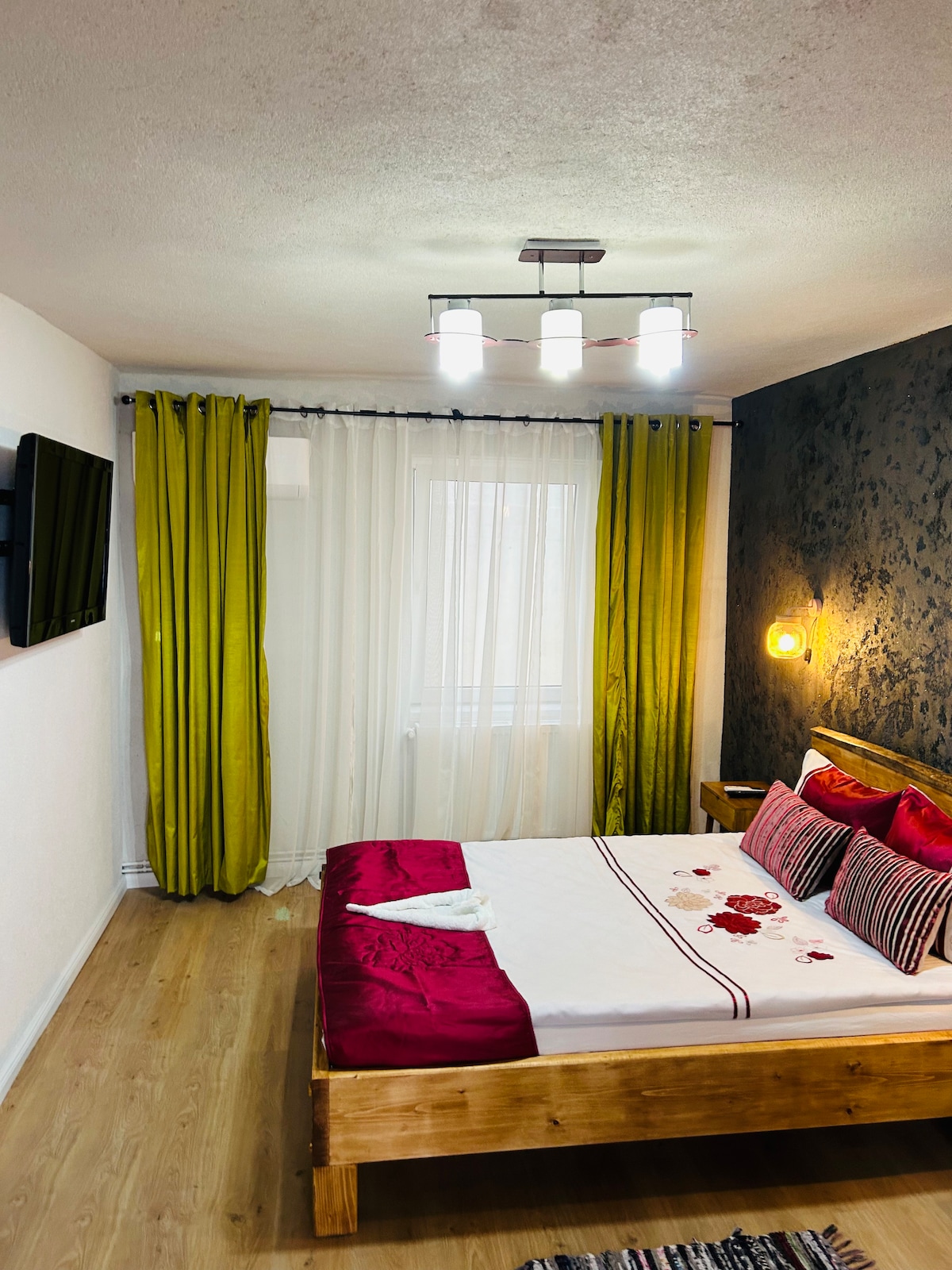 A spacious bedroom is featured with a double bed adorned with decorative pillows and a blanket. Large windows are framed with bright green curtains, allowing natural light to fill the room. A television is mounted on the wall, enhancing the modern design.