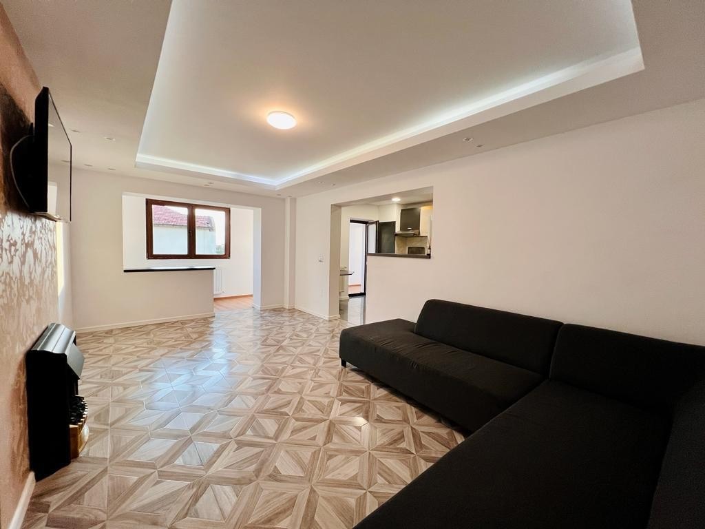 An open living area features a modern black sectional sofa positioned on patterned tile flooring. Natural light enters through a large window, illuminating the space, while a kitchenette is visible in the background, showcasing a simple and functional layout.