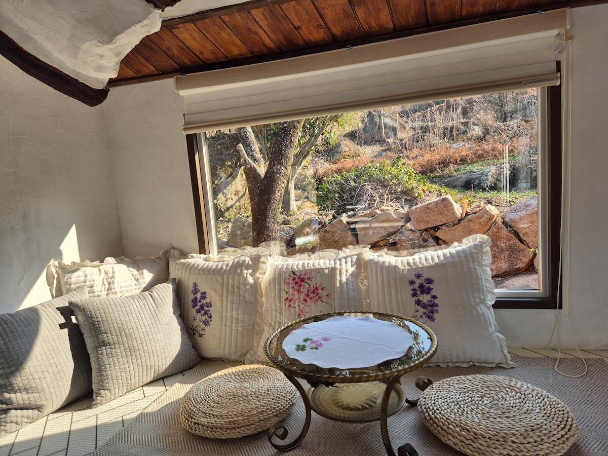 A cozy seating area features a low glass table surrounded by soft cushions with floral patterns. Natural light pours in through a large window, offering a view of the serene outdoors with trees and rocky terrain.