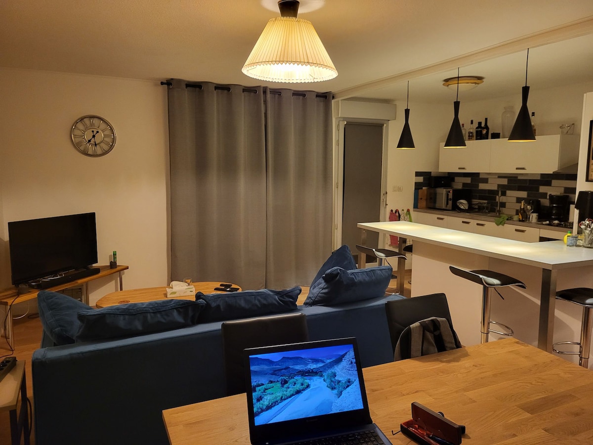 A modern living area features a blue sofa facing a television, with a wooden dining table in the foreground. Pendant lights hang above a bar-style kitchen counter, which displays a sleek design with dark and light accents. Gray curtains are partially drawn.