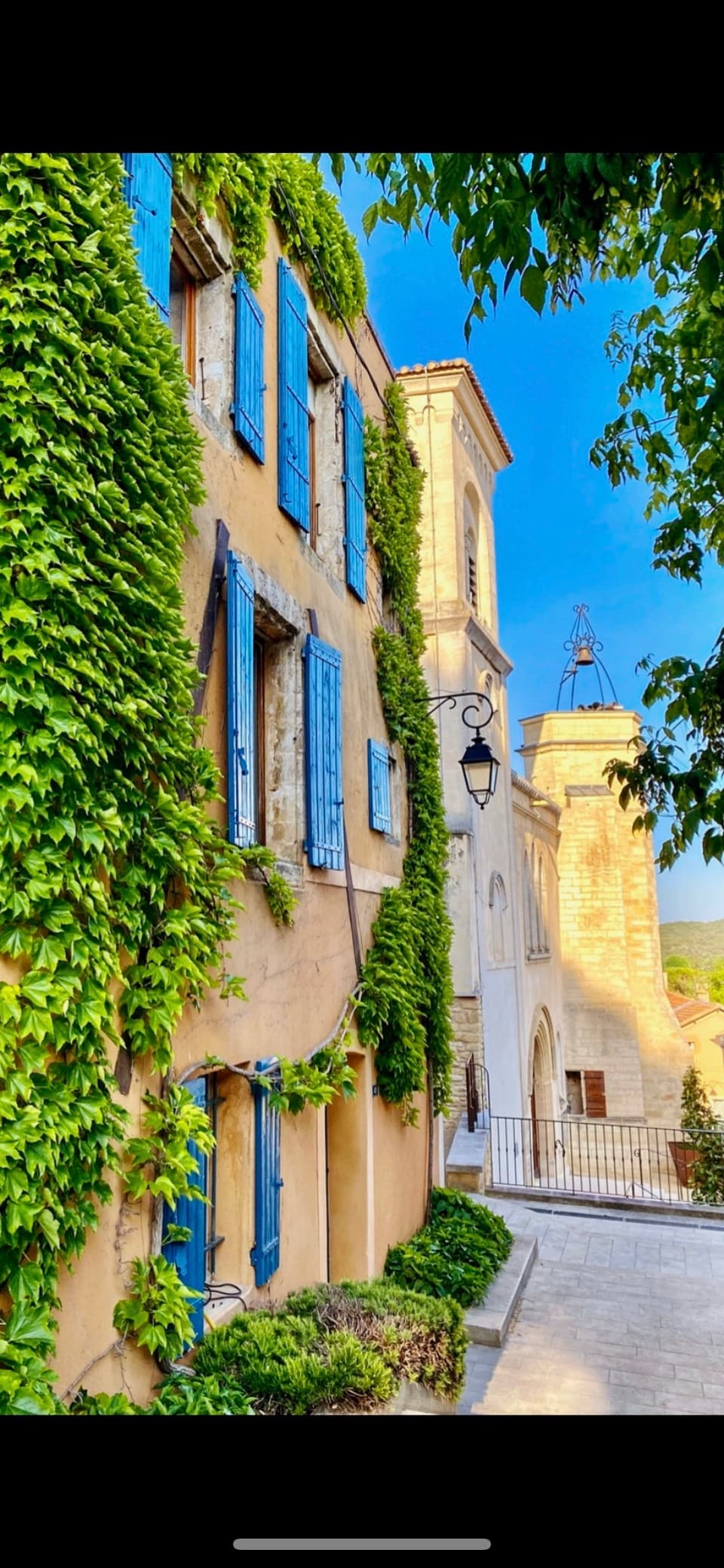 A charming stone building displays blue shutters and is adorned with green ivy. The structure features a visible clock tower in the background against a clear blue sky. Cobblestone pathways lead around the buildings, creating an inviting and quaint atmosphere.