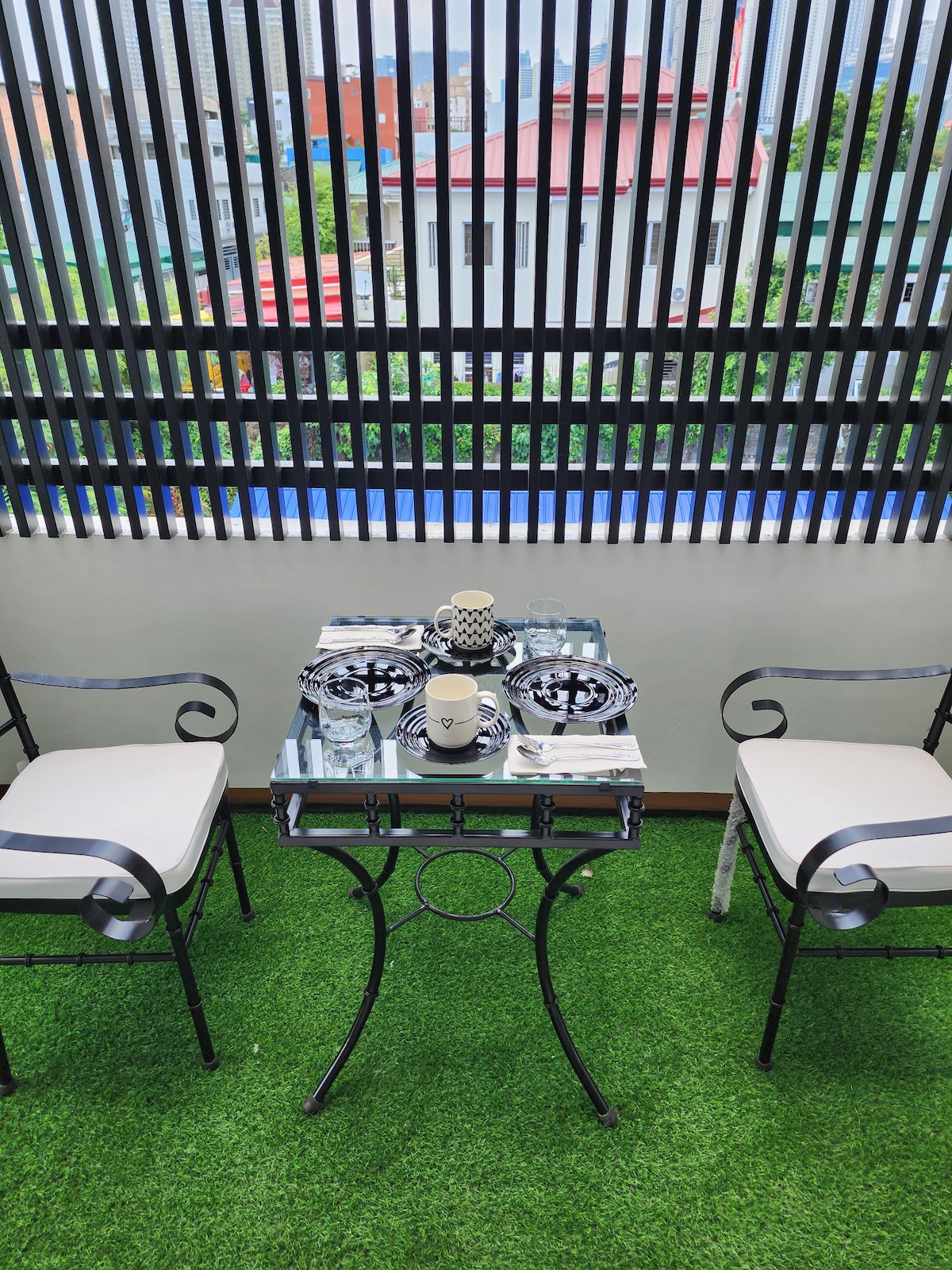 A small outdoor dining table is positioned on a green turf surface, accompanied by two chairs. The table is set with dinnerware and candles, while a patterned tablecloth adds a decorative touch. Vertical slatted panels provide partial privacy and an urban view.