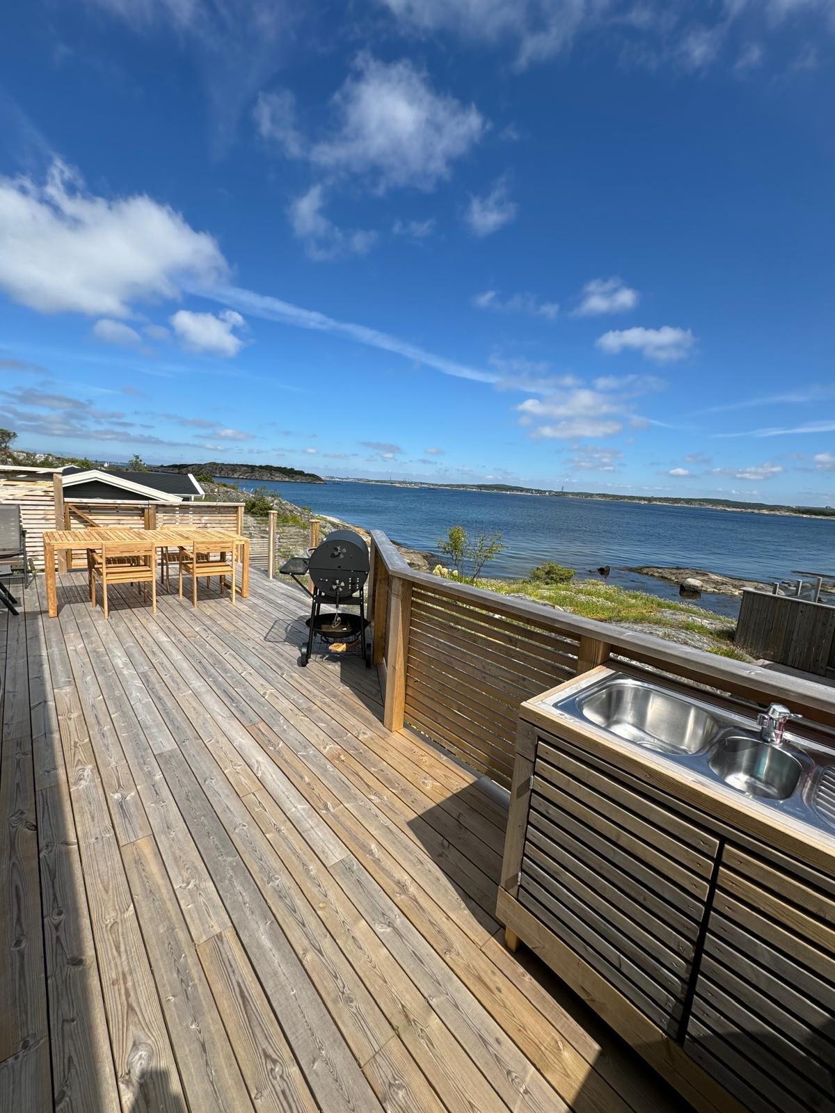 A spacious wooden deck overlooks the tranquil sea, featuring a wooden dining table with seating for six, a barbeque grill, and an outdoor kitchen area. The deck offers unobstructed views of the water and nearby rocky shore, framed by a clear blue sky.