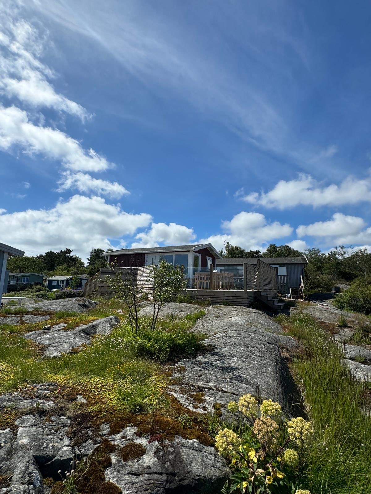 A cozy stuga is nestled among natural rock formations, framed by a vibrant green landscape. The building features large windows that invite ample daylight, and the sky above is filled with scattered clouds, hinting at a pleasant day outdoors.