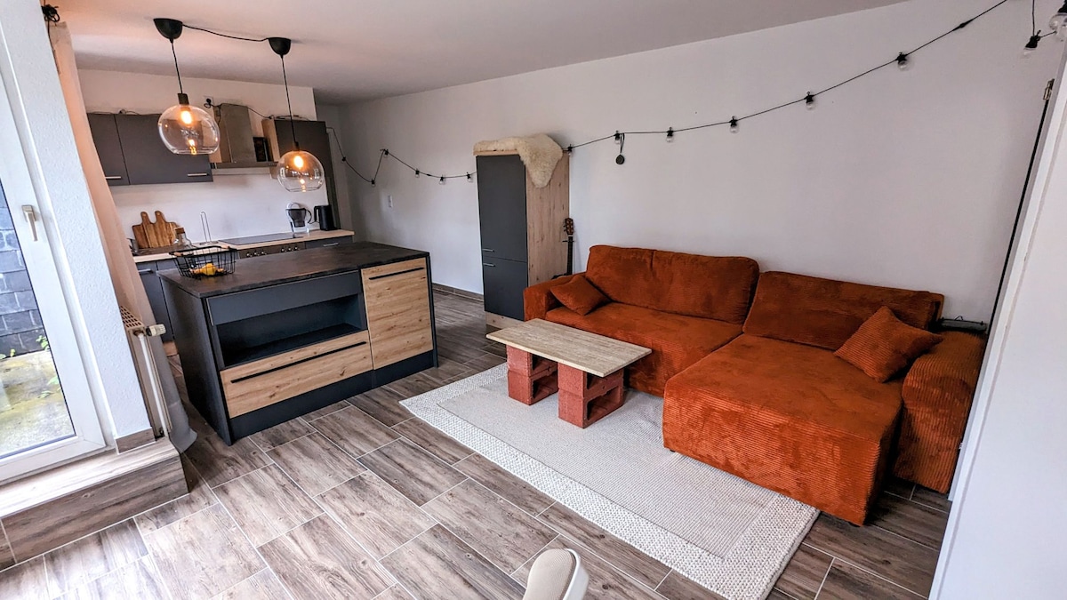 A contemporary living space features an orange sofa and matching cushions, positioned near a wooden coffee table. A modern kitchen area is visible, complemented by stylish pendant lights. Wooden flooring extends throughout, while string lights add a subtle ambiance to the room.
