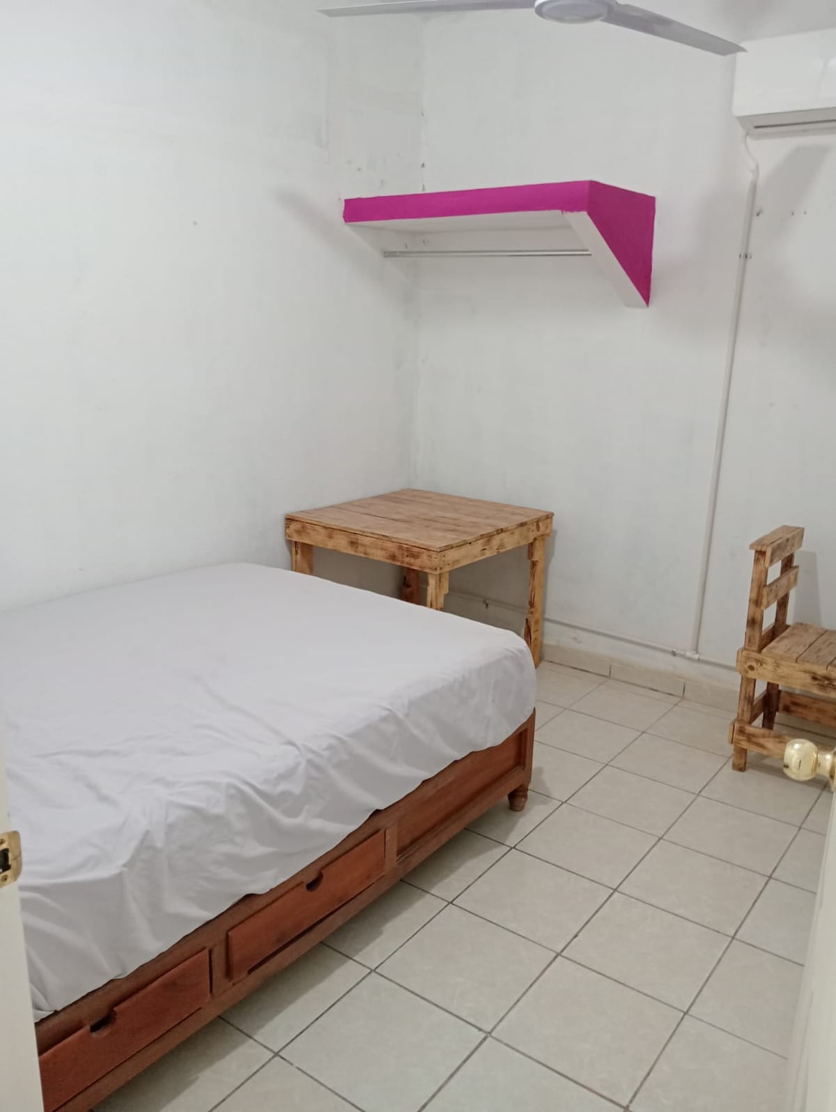A simple room features a spacious bed with a white coverlet, positioned against a plain white wall. A wooden table and a chair are situated nearby. A shelf in a vibrant color adds a touch of contrast above the table. The floor is tiled.