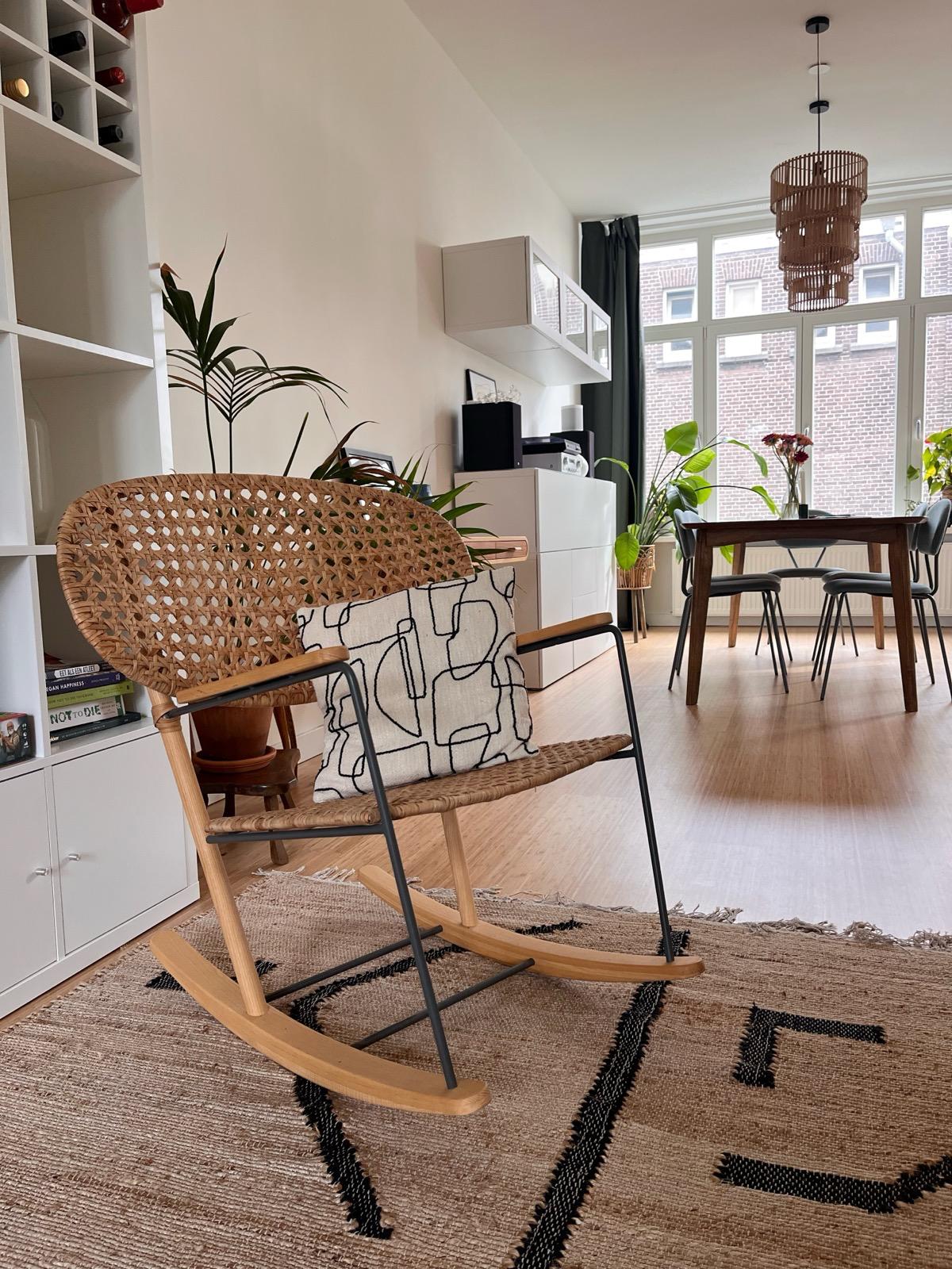A woven rocking chair is positioned in a well-lit living area, which features a mixture of greenery and natural light streaming through large windows. In the background, a dining table with chairs and additional plants are visible, enhancing the space's warm and inviting feel.