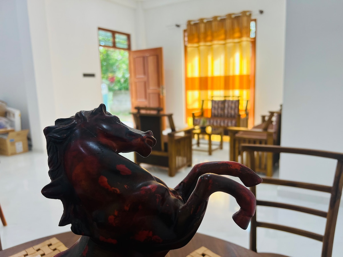 A detailed view of a decorative horse figurine is captured in the foreground, with a living area visible in the background. The space features wooden seating arrangements and large windows adorned with sheer orange curtains, allowing natural light to filter through.