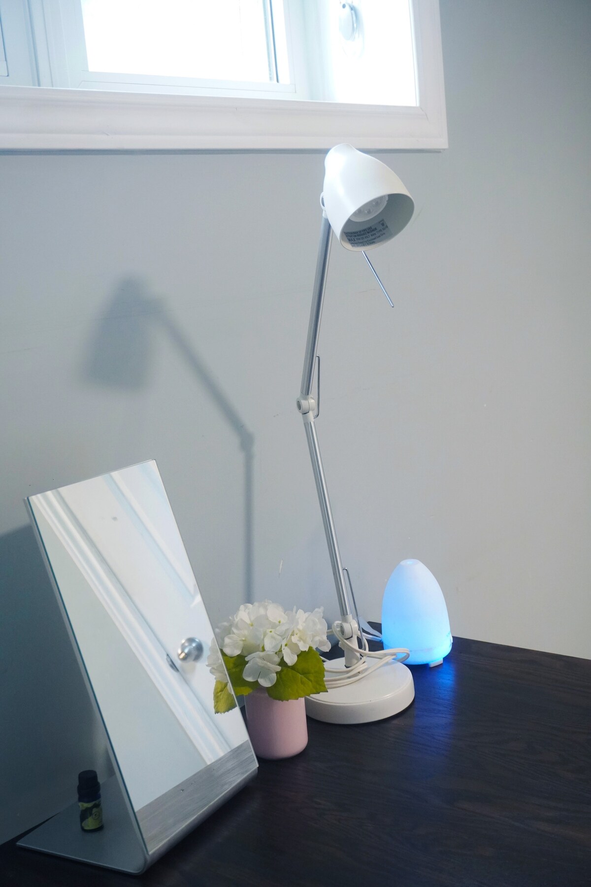 A desk area features a sleek, adjustable lamp casting soft light alongside a modern mirror. A small pink vase holds white flowers, and a gentle blue light source adds a calming touch to the arrangement.