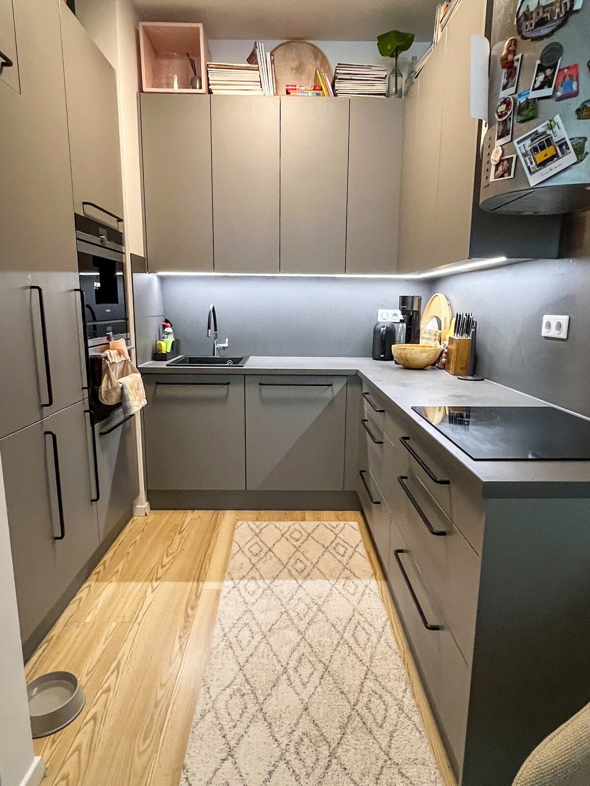 A modern kitchen features sleek cabinets and countertops in a muted color palette. Under-cabinet lighting highlights the workspace. A sink and cooking area are positioned near the back, with a patterned rug covering the floor. Magnetic decorative items are present on the refrigerator.