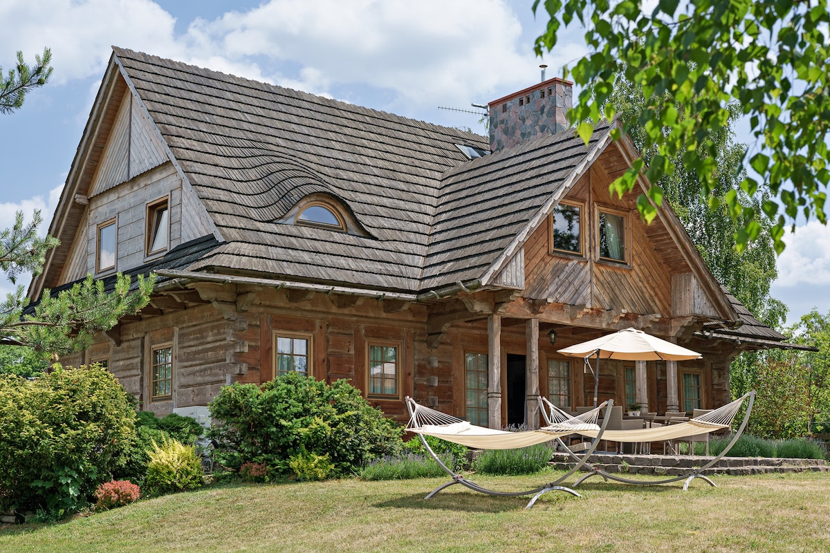 A charming wooden house features a steeply pitched roof and several large windows. Two hammocks are positioned on the lawn, accompanied by an umbrella for shade. Lush greenery surrounds the structure, creating a serene outdoor environment.