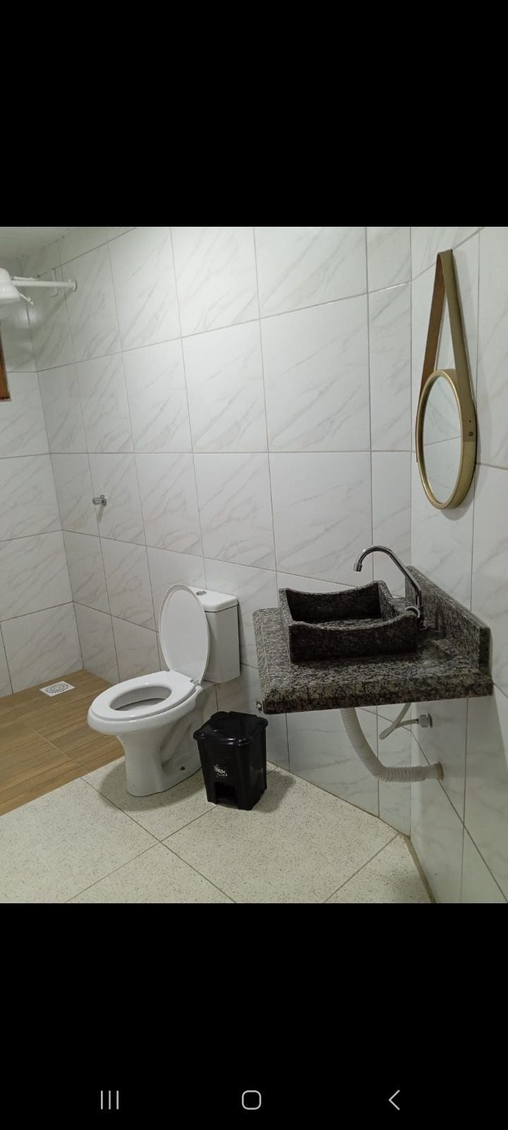 The bathroom features a modern design with white tiled walls and a spacious granite countertop. Two sinks are positioned side by side, complemented by a round mirror above. A toilet is located nearby, and a small black wastebasket is placed on the floor.