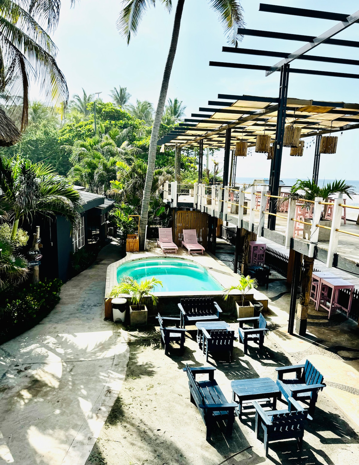 An inviting outdoor space is highlighted by a turquoise pool surrounded by lounge chairs. Lush greenery and tall palm trees provide shade, while the elevated structure features a shaded area with hanging light fixtures above. The area appears spacious and well-designed for relaxation.