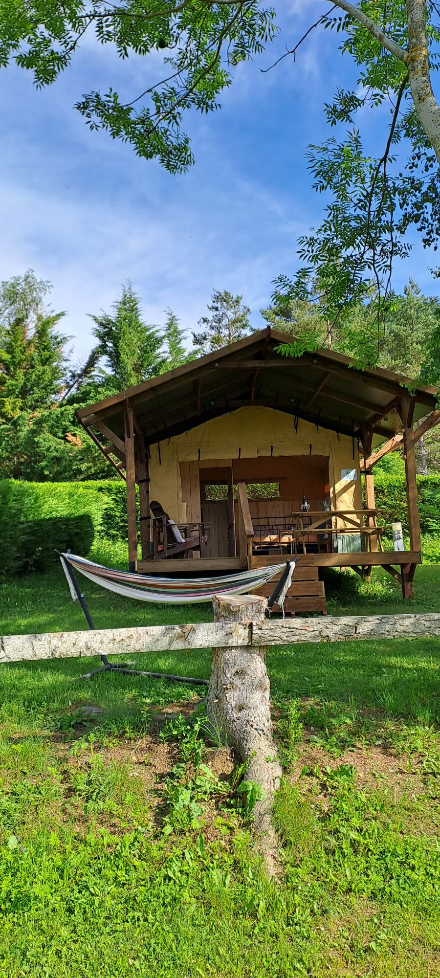 A charming safari tent is set on a grassy area, surrounded by lush greenery. A covered porch is featured at the front, with a hammock hanging nearby. The structure’s wooden beams provide a rustic touch, inviting relaxation in a serene outdoor setting.