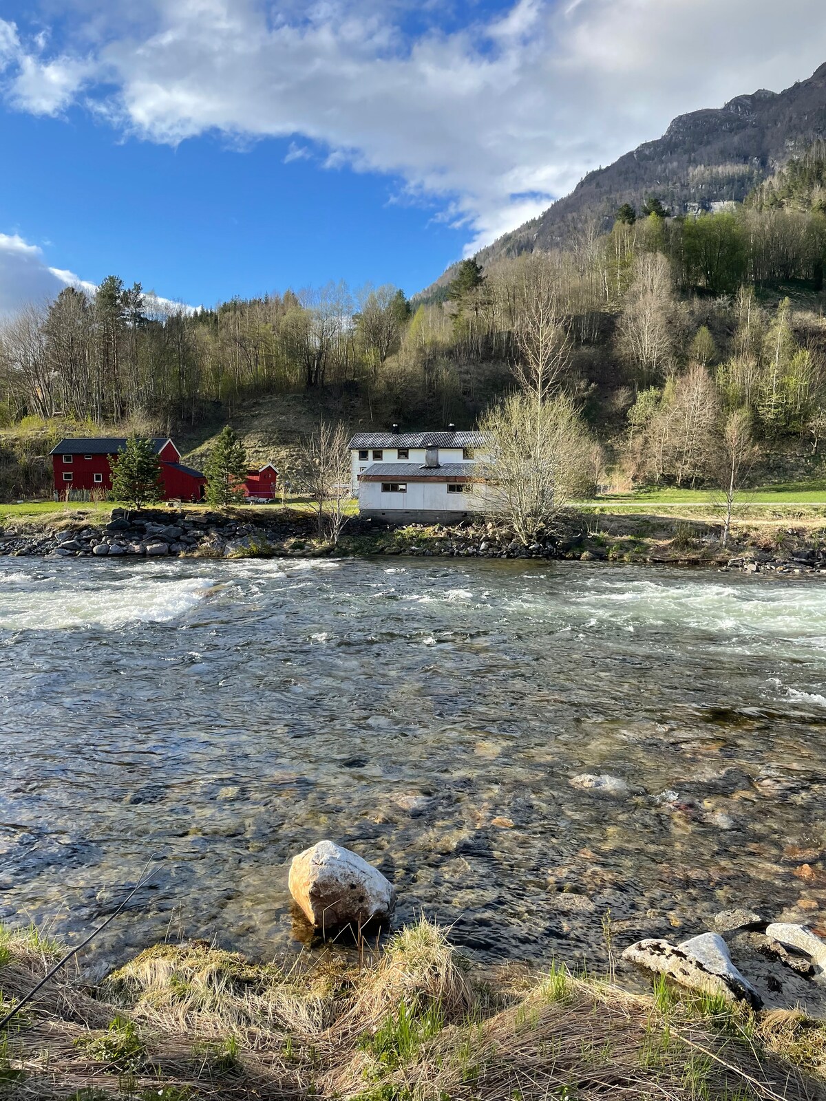 The image showcases a tranquil riverside view with a meandering river in the foreground. A charming white house and two red outbuildings are situated against a backdrop of lush greenery and mountains, under a partly cloudy sky. The scene highlights the natural beauty of the surroundings.