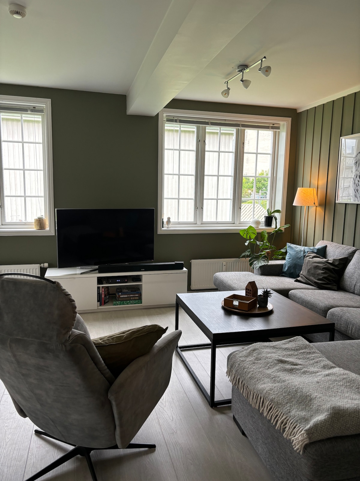 A cozy living area is defined by a gray sectional sofa and a comfortable armchair. A coffee table stands in the center, with a decorative tray placed on top. Natural light filters through two large windows, complemented by soft lighting from a nearby lamp.