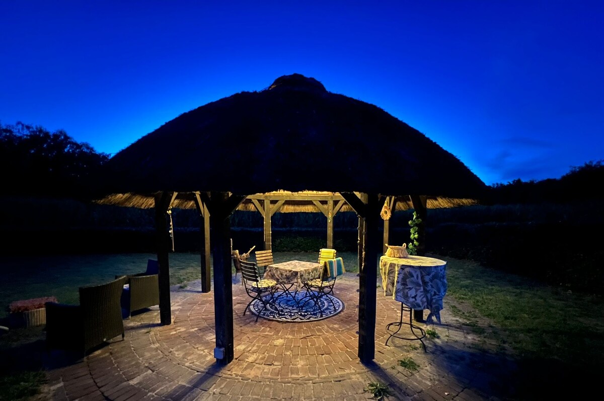 An outdoor gazebo is illuminated under a twilight sky, showcasing a circular seating area with wooden beams and a thatched roof. A round table is positioned in the center surrounded by chairs, while a lush garden surrounds the structure, enhancing the serene outdoor setting.