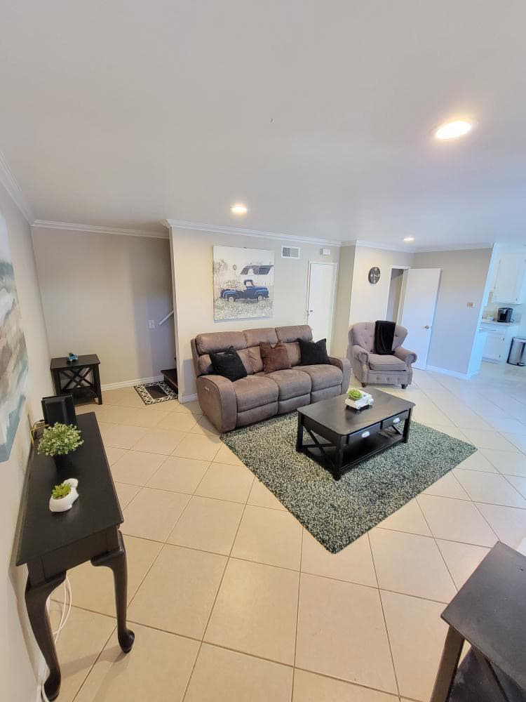 A spacious living area features a reclining three-seat couch facing a coffee table on a textured rug. Two accent chairs sit nearby, and a small table holds a plant. Light beige walls and tile flooring create a warm ambiance, complemented by a decorative wall art piece.