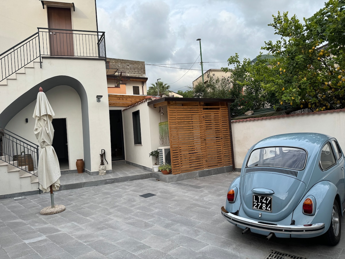 An outdoor area is presented with a grey tiled surface, featuring a vintage blue car parked nearby. A light-colored umbrella stands nearby, and a wooden structure with slatted design is visible in the background, accompanied by greenery.