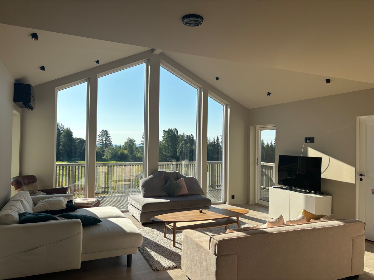 A spacious living area is illuminated by natural light through large triangular windows. Comfortable seating includes a sectional sofa and individual chairs, arranged around a central coffee table. A flat-screen television is positioned near the entrance to a balcony, enhancing the indoor-outdoor connection.