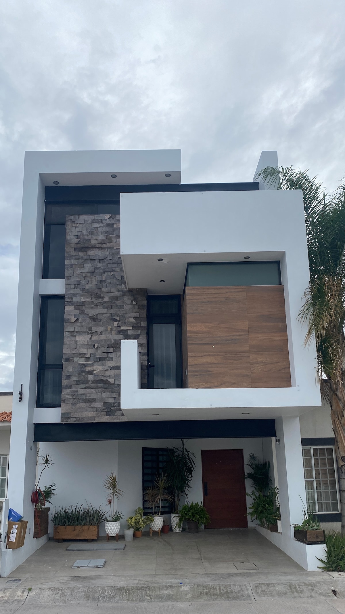 The modern three-story building features a combination of stone and wood accents on the facade. Large windows allow for natural light, while potted plants add greenery to the entrance area. A spacious porch leads to a wooden front door.