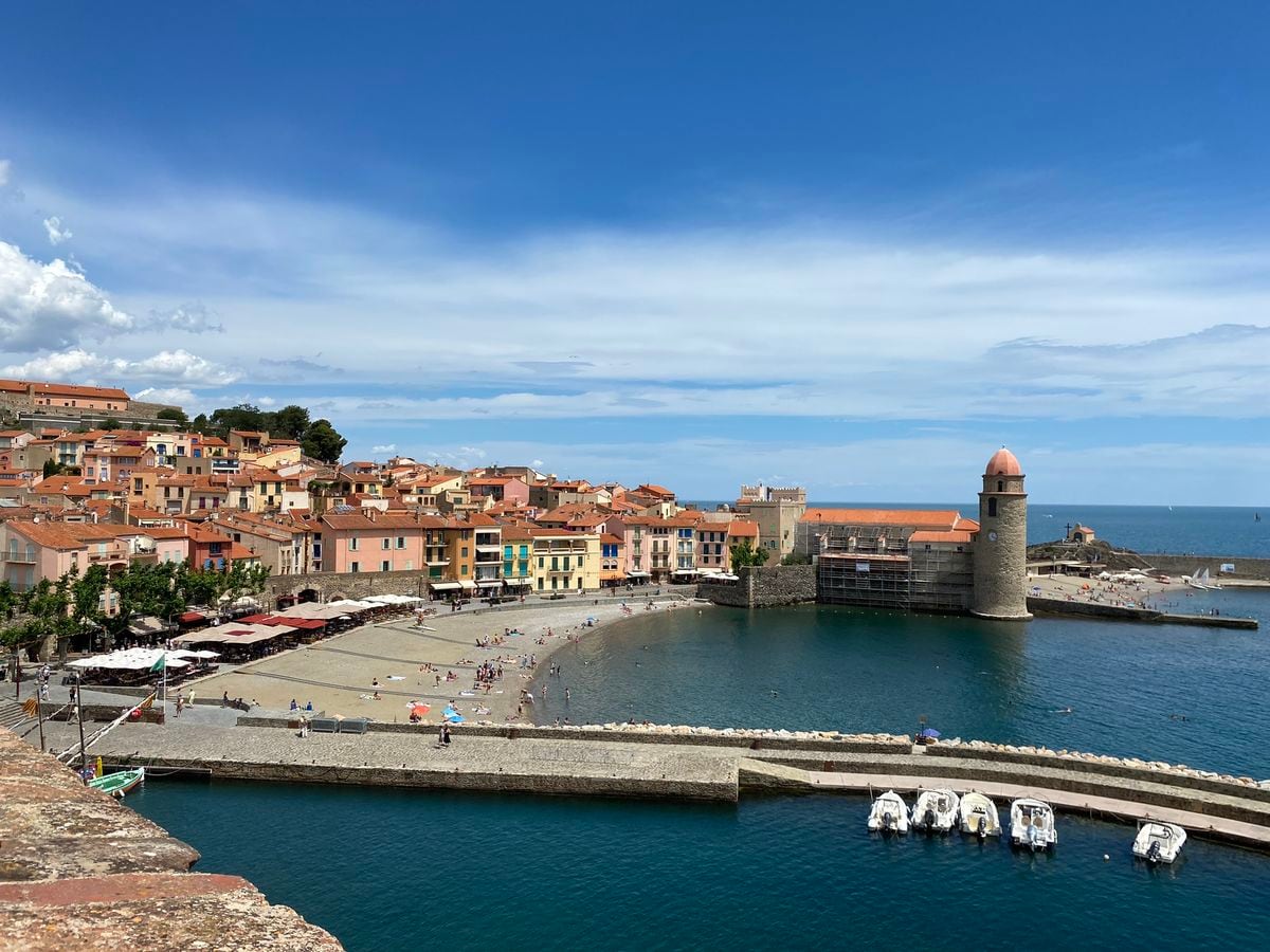 A coastal view captures the vibrant colors of the village buildings lining the beach. The calm water reflects the lively scene, with small boats docked nearby. A historic structure with a rounded tower stands along the shore, providing a scenic focal point against a clear blue sky.