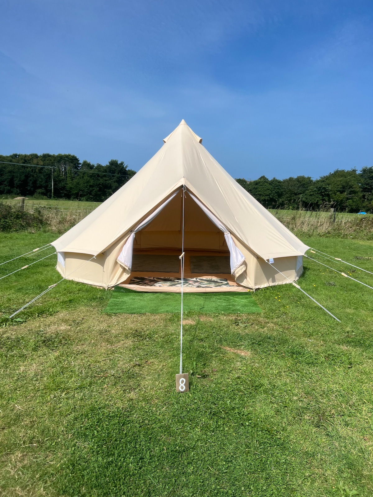 A light-colored bell tent is positioned on a grassy area, featuring an open entrance that invites natural light. The surrounding landscape includes lush greenery and a clear blue sky, enhancing the outdoor camping experience.