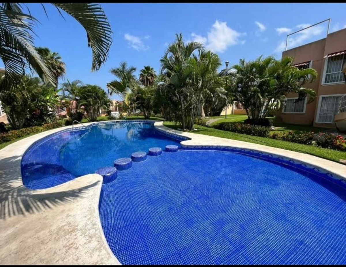 A large swimming pool is framed by tropical plants and well-maintained landscaping. The pool features a unique shape with steps leading into the water, showcasing bright blue tiles that reflect sunlight under a clear sky.