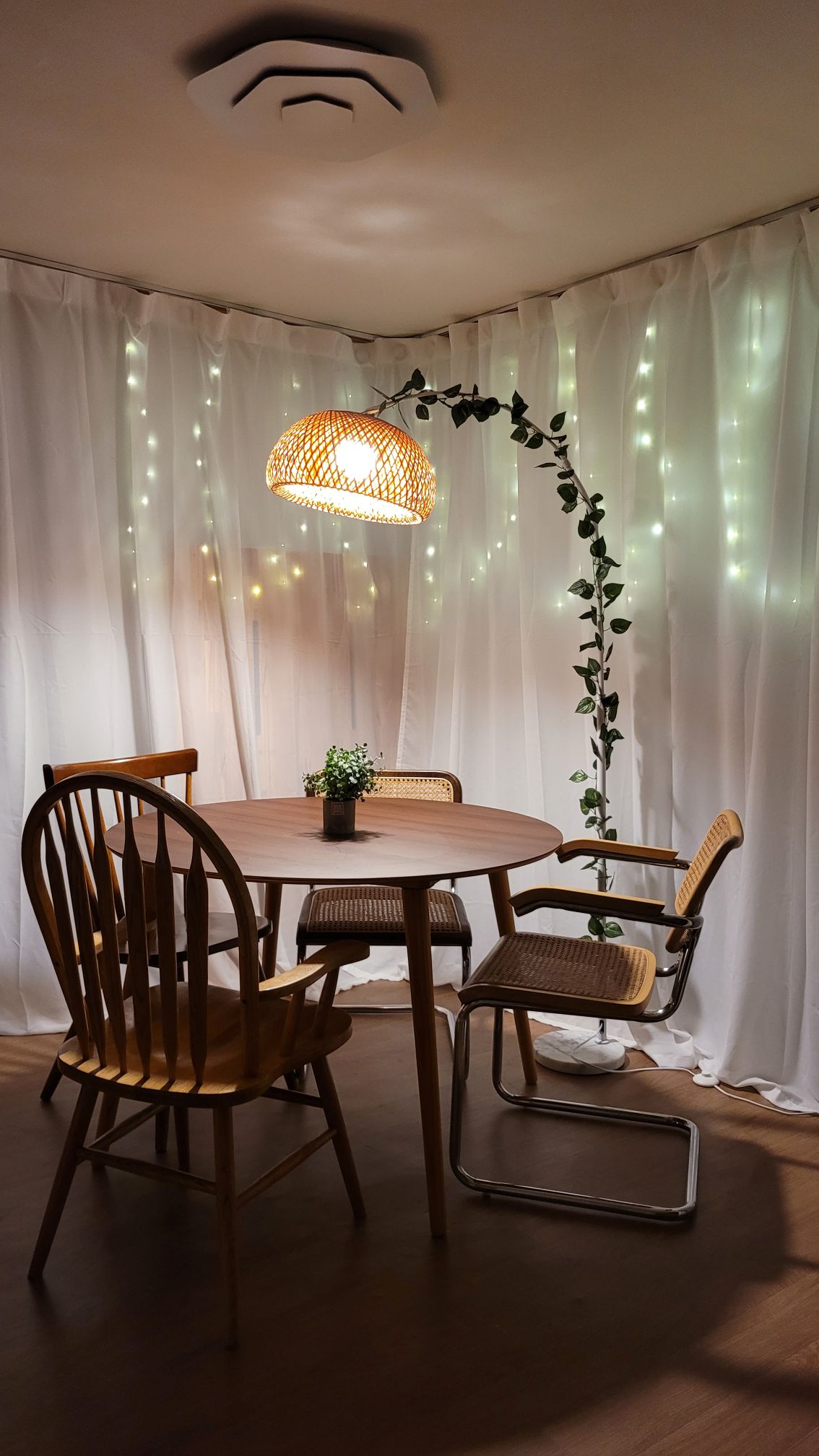 A dining space is highlighted by a round wooden table surrounded by four chairs, including two wooden ones and two with metal frames. A soft glow from a pendant lamp and strings of lights create a warm atmosphere, complemented by a small potted plant on the table.