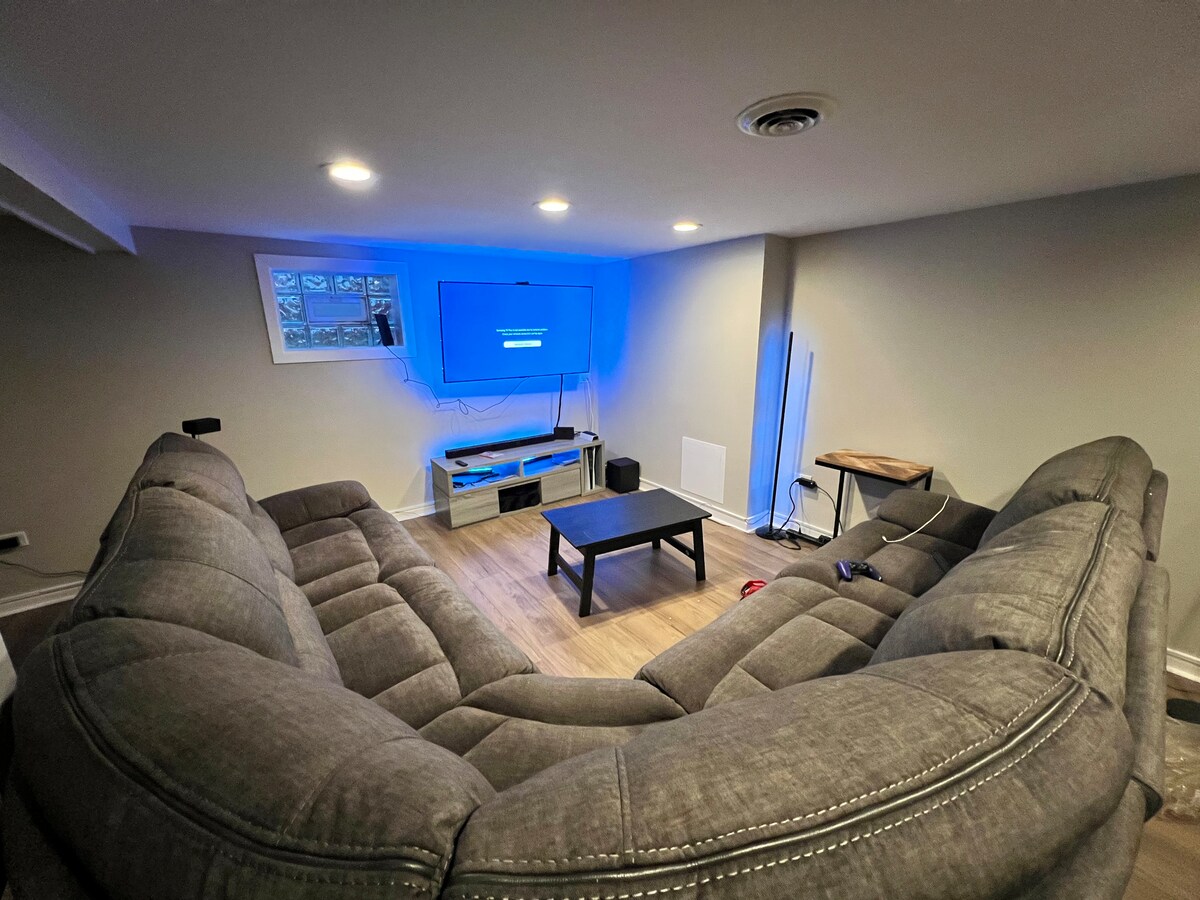 The living room features a spacious sectional couch arranged to face a mounted TV, which is enhanced by LED lights that change colors. A coffee table sits centrally, and additional seating options are available. Natural light filters through small windows above.