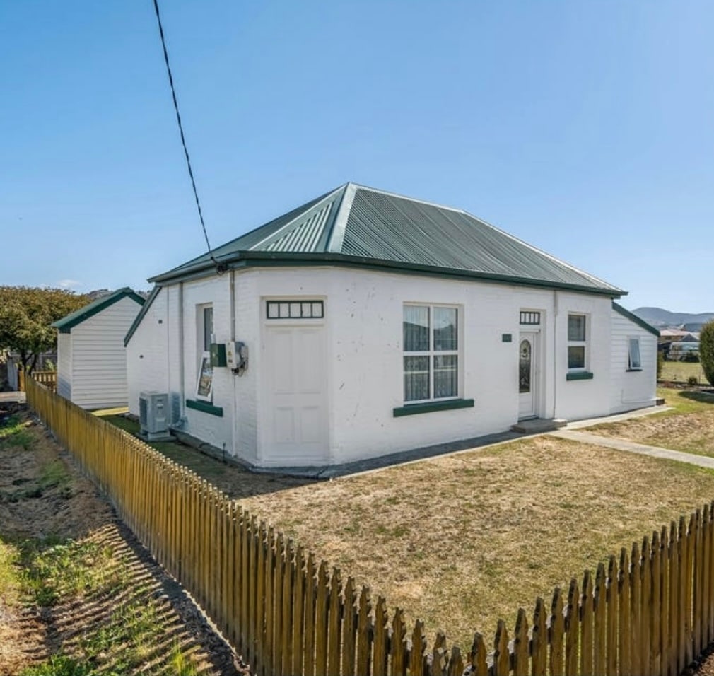 The heritage cottage features a classic white exterior complemented by green trim. A well-maintained lawn surrounds the home, and a wooden picket fence provides a charming boundary. Clear skies above enhance the inviting rural setting.