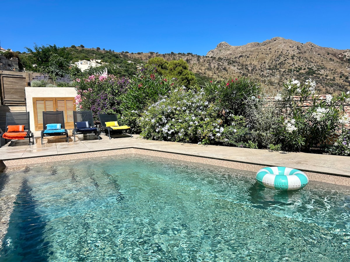 A tranquil pool area is surrounded by lush greenery and colorful flowers. Several lounge chairs are positioned around the unheated pool, inviting relaxation. A buoy floats gently in the water, and the mountains are visible in the background under a clear blue sky.