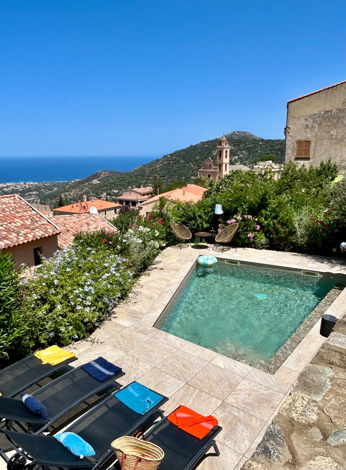 A private pool is surrounded by a stone terrace, with colorful sun loungers positioned nearby. Lush greenery and blooming flowers line the edge, providing a serene backdrop. In the distance, sea views and mountain scenery are visible under a clear blue sky.