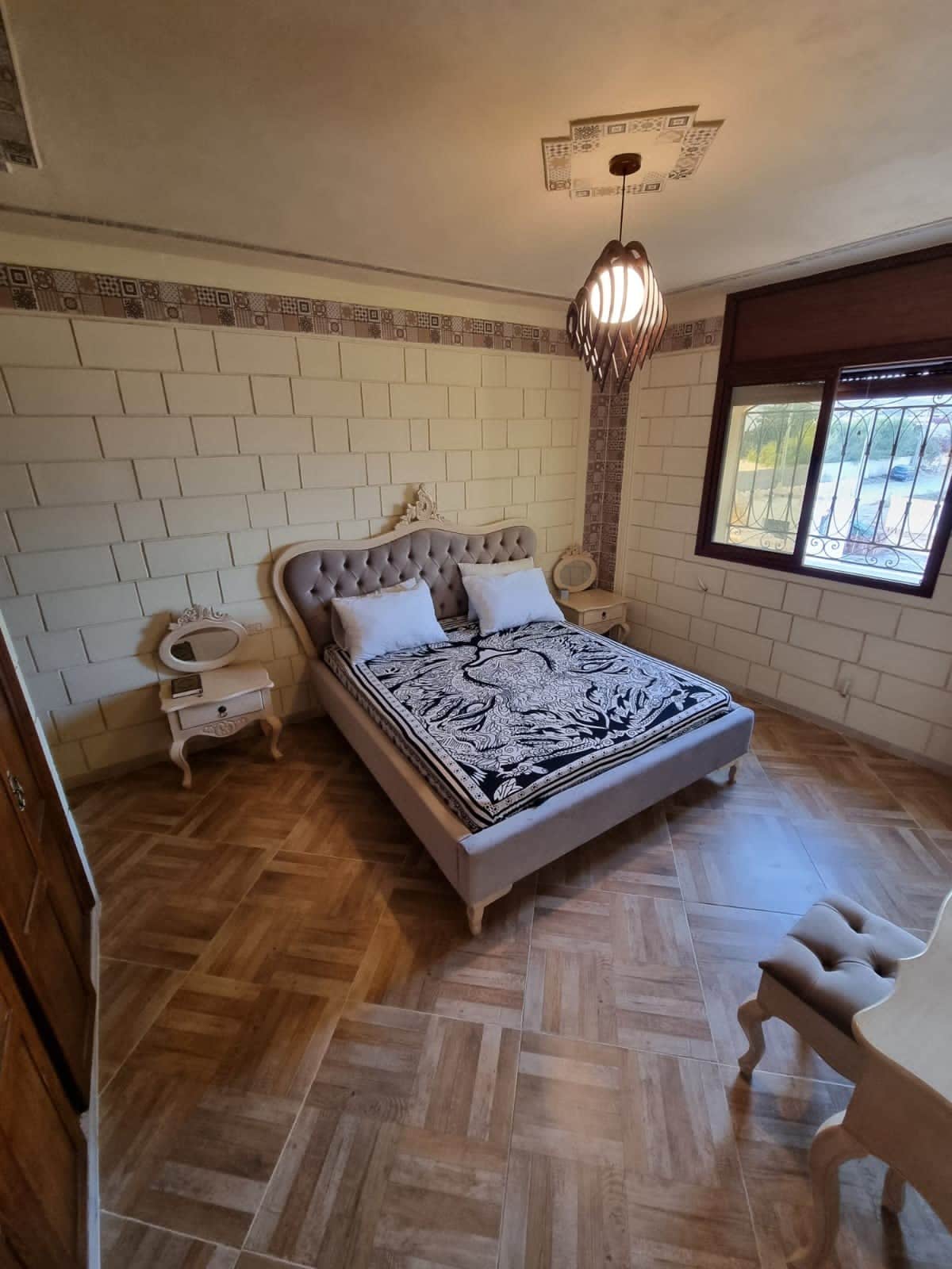 A cozy bedroom features a spacious upholstered bed adorned with patterned linens. Soft light filters through a large window with a security grate, illuminating the hardwood floor. Decorative bedside tables are positioned on either side of the bed, enhancing the inviting decor.