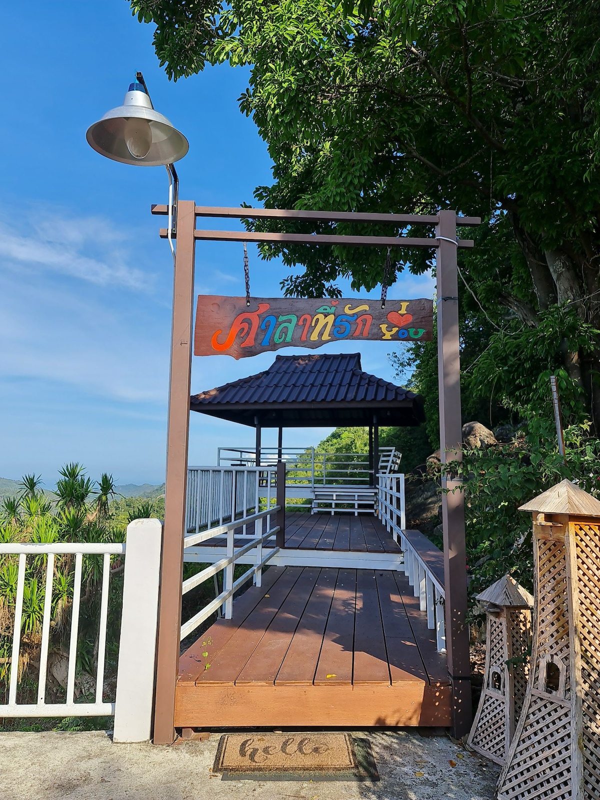 A welcoming entrance is framed by a wooden structure featuring a sign that reads 'You' in colorful letters. The path leads to a shaded gazebo with a traditional roof, surrounded by greenery, with a cozy mat welcoming guests at the foot of the steps.